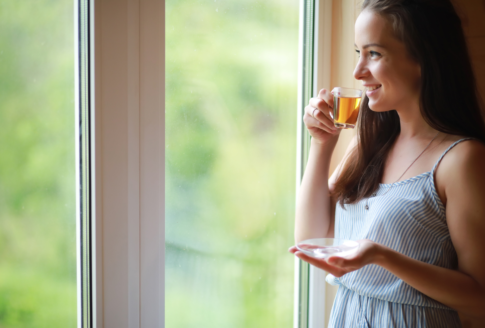 young woman drinks tea upright next to window