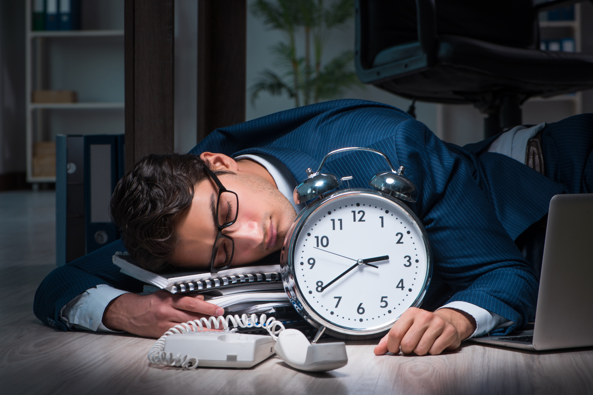 άντρας με μπλε κουστούμι κοιμάται στο γραφείο του δίπλα σε ρολόι