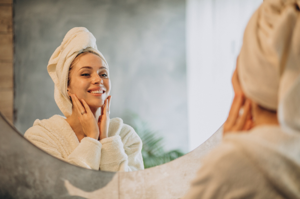γυναίκα με μπουρνούζι & πετσέτα στο κεφάλι κοιτά στον καθρέφτη το δέρμα της
