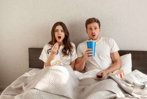 ζευγάρι κάθεται στο κρεβάτι τρώγοντας & πίνοντας μπροστά στην τηλεόραση